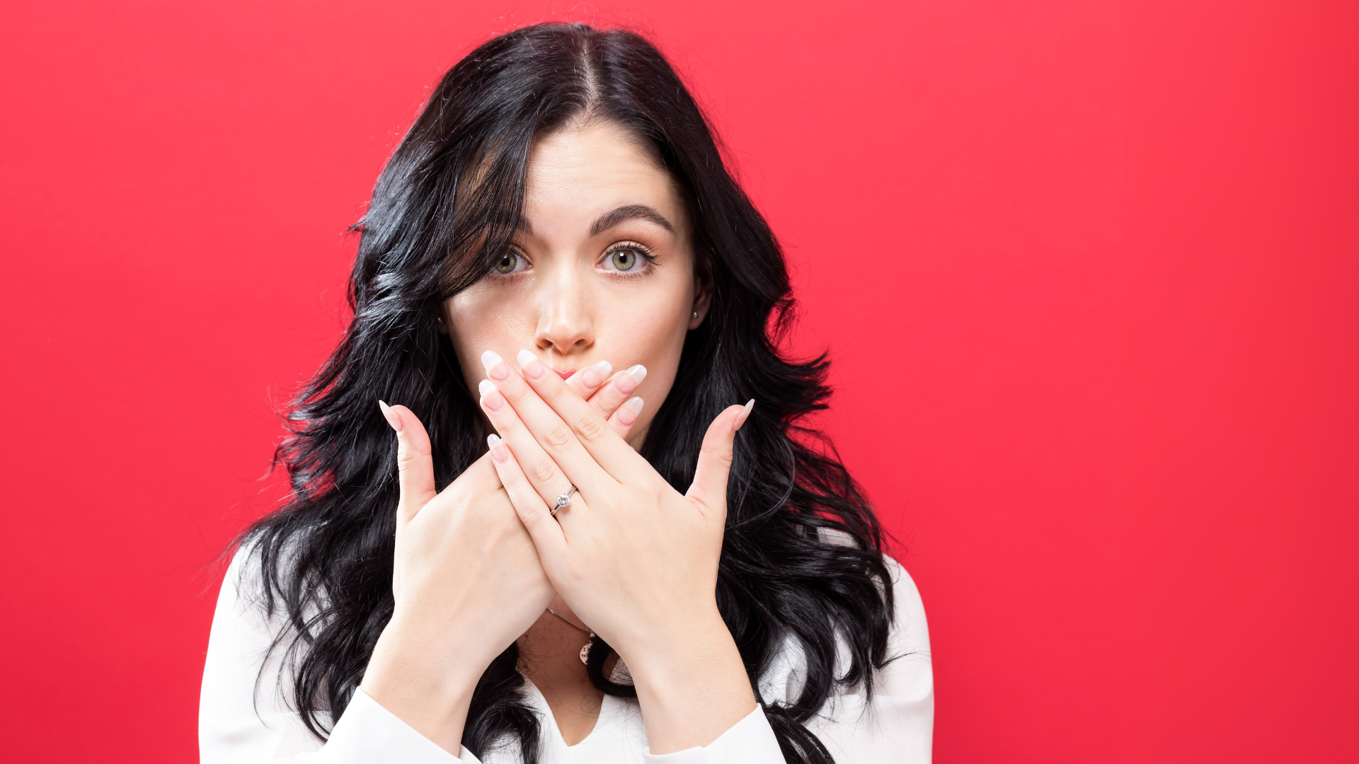 A young woman with long, dark, wavy hair and light eyes looks directly at the viewer with a surprised expression. She is covering her mouth with both hands, her fingers interlocked. She is wearing a white top and a ring is visible on her left ring finger. The background is a solid red colour.