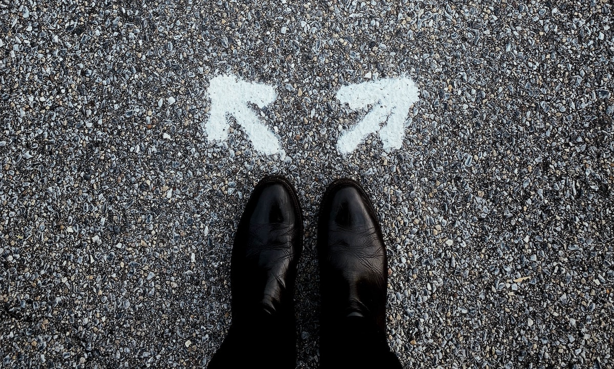 two feet encased in shiny black shoes stand in front of two arrows spray painted on the road, pointing in different directions.