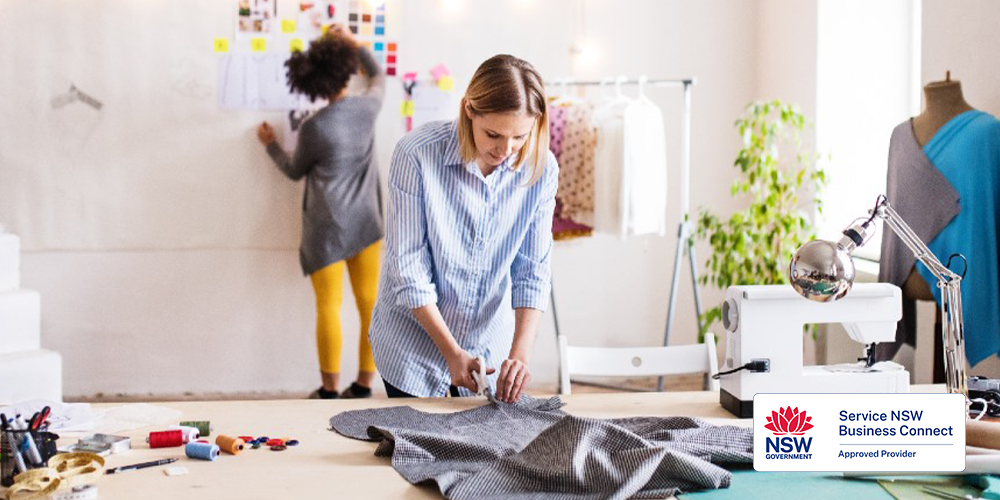 In a well-lit and creative studio space, a woman is focused on cutting fabric laid out on a large work table. The table is scattered with various sewing tools and materials. In the background, another woman is working on a design board, pinning up sketches and fabric swatches. The space is decorated with plants and a rack of clothing, suggesting an environment dedicated to fashion design or crafting. In the bottom right corner, there is a logo for Service NSW Business Connect, indicating an approved provider.