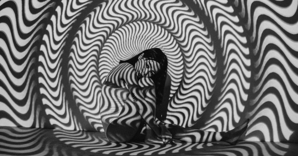 A woman sits on the floor, her body curled up with her arms wrapped around her knees. She is surrounded by a dynamic black and white spiral pattern, likely projected onto her and the walls. The swirling lines create an optical illusion effect, casting shadows and light across her figure. The image has a surreal and hypnotic quality, blending the human form with abstract visual elements