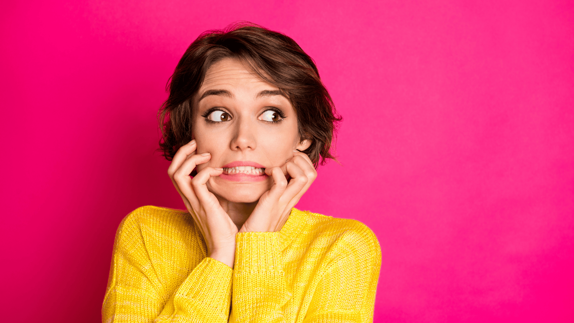 A young woman with short brown hair wearing a bright yellow sweater stands against a vibrant pink background. She has a nervous or anxious expression, biting her lower lip and gripping her fingers near her mouth. Her wide eyes look to the side, conveying uncertainty or worry about funding application mistakes.