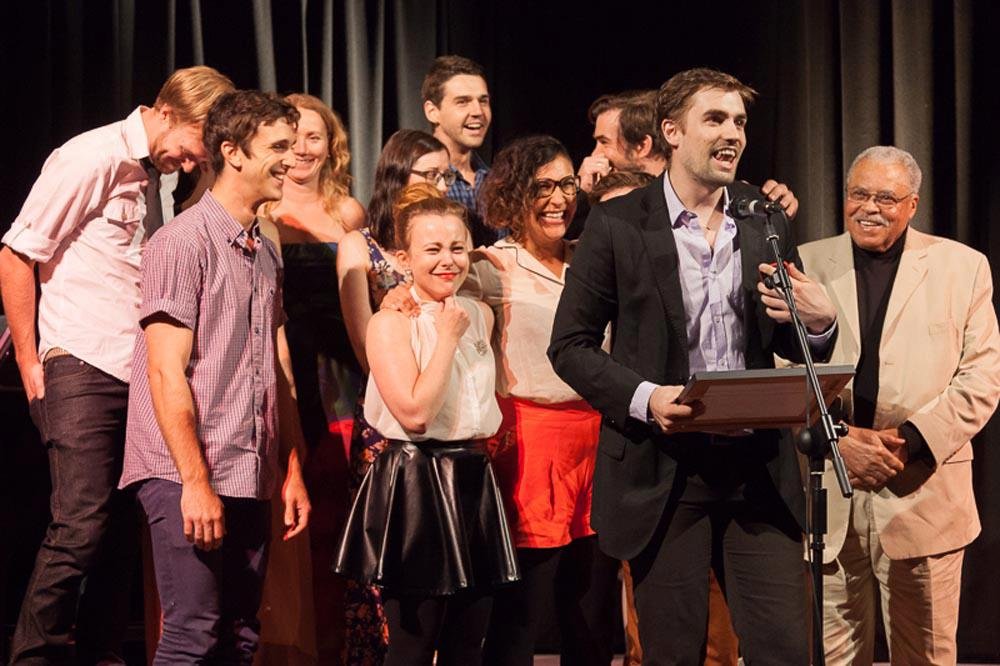 A group of about ten people are on a stage, some standing close together, others with arms around each other. One man in a dark suit speaks into a microphone, holding an award. They are celebrating, and many are smiling. The background is a dark stage curtain. The lighting is bright, illuminating the group. On the far right, an older gentleman in a light-colored suit stands with his hands clasped. The group appears to be diverse and of varying ages.