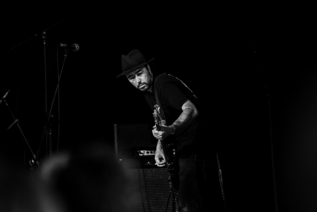 A black and white photo shows Sats Kramer on stage playing a guitar, wearing a fedora and a dark t-shirt. He is looking down at the strings of his guitar, which he holds with both hands. His left arm reveals several tattoos. In the background, a speaker cabinet is visible, and microphones are scattered around the dimly lit stage.