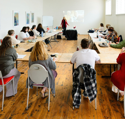 Workshops A group of people seated at tables facing a speaker. They are attending a creative business workshop delivered by Creative Plus Business.