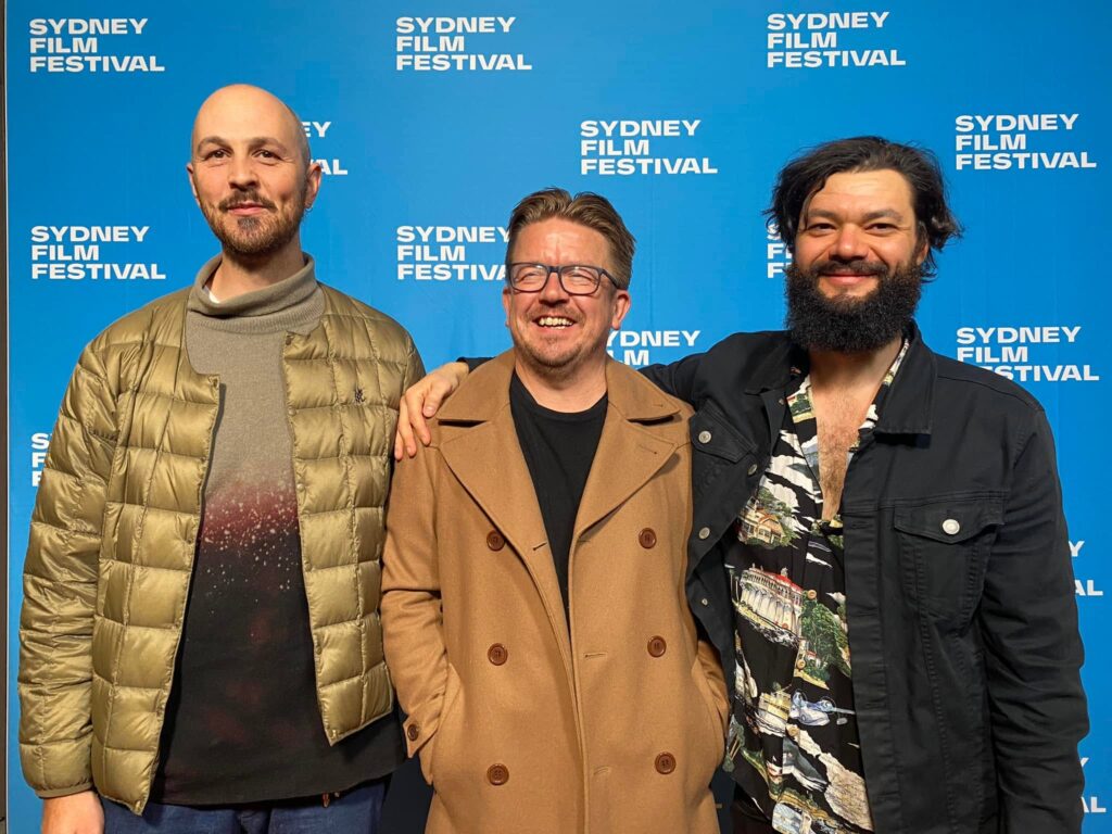 Three men stand side-by-side in front of a blue wall with the "Sydney Film Festival" logo repeated in white. The man on the left has a shaved head and a beard, wearing a gold puffer jacket over a grey and red-patterned turtleneck. The man in the middle has glasses and short brown hair, a beard, and a brown double-breasted coat over a black shirt. He has his left arm around the shoulder of the man on the right. The man on the right has dark, curly hair and a beard, wearing a black jacket over a patterned shirt. All three men are smiling.