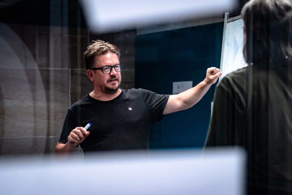 A man with short brown hair, a beard, and glasses, wearing a black t-shirt, stands in front of a dark background, looking intently at someone off-camera to his right. He holds a blue marker in his left hand and has his right arm raised and bent at the elbow, with his fist clenched, as if making a point or demonstrating something. The image is taken through a reflective surface, with out-of-focus white shapes in the foreground.