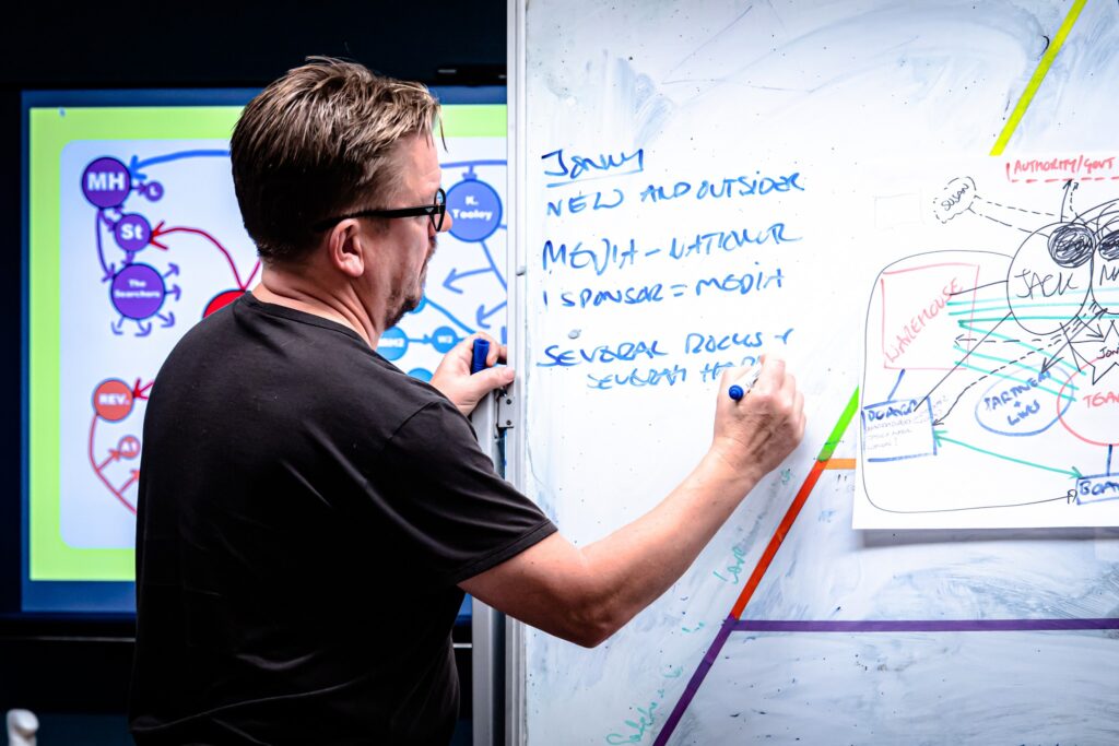 A man with short, light brown hair and glasses, seen from behind and to the left, is writing on a large white board with a blue marker. He is wearing a black t-shirt. On the whiteboard, there is handwritten text in blue marker, and some diagrams, one of which includes the words "JACK." To the left of the man, a screen displays a colourful diagram with various interconnected circles and text.