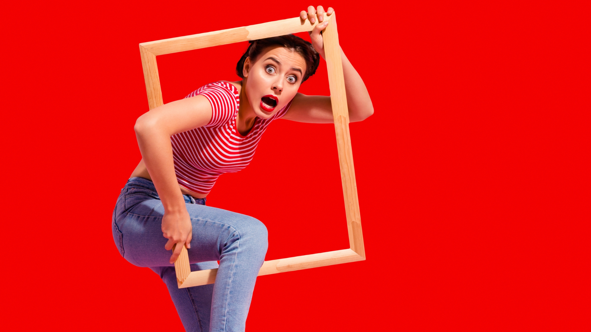 A surprised-looking woman in a red and white striped t-shirt and jeans is climbing out of or escaping through a wooden picture frame, with a dramatic expression. The background is solid bright red.