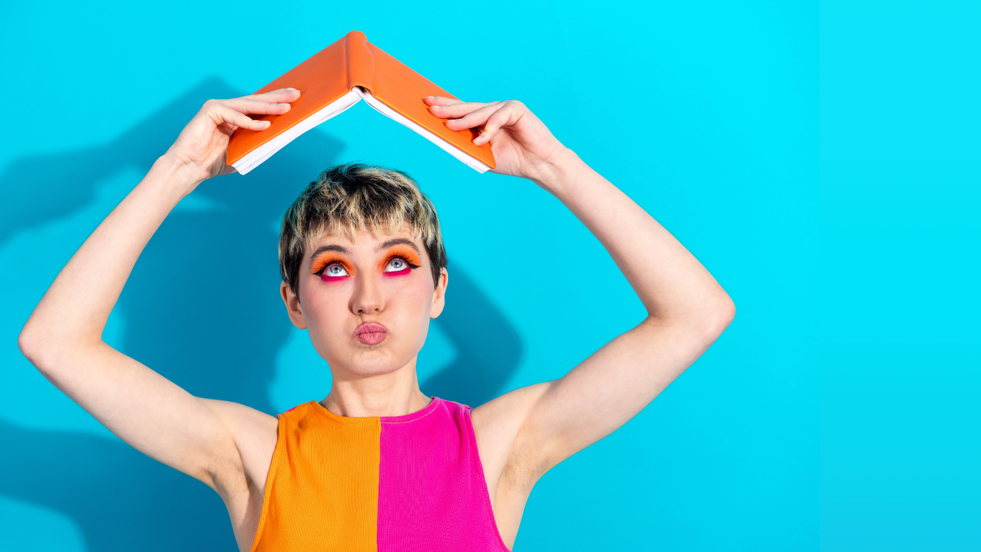 Blog Hero Image for Learning to Thrive A vibrant, high-contrast photo of a person with short blonde and brown hair centred against a bright cyan blue background. They are wearing an orange and pink sleeveless top and bold orange eye makeup, holding an open orange book over their head like a roof. They have a playful, pouting expression while looking upward toward the book.