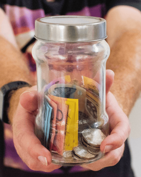Small hero image for the cashflow for creatives workshop. A close-up of a person's hands holding a clear glass jar filled with a mix of Australian banknotes and various silver and gold coins.