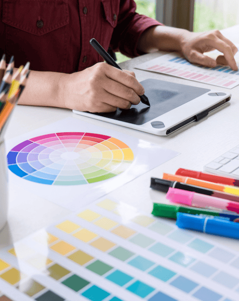 Small hero image for the creative's guide to branding workshop. A person in a maroon shirt works at a bright white desk, using a digital drawing tablet and stylus. The workspace is cluttered with creative tools, including several colour wheels, printed colour swatches, a jar full of coloured pencils, and a set of vibrant markers