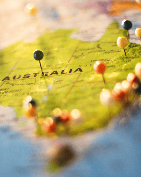 Small promo image for the Cultural Tourism Workshop. A close-up, angled shot of a map of Australia with the name "AUSTRALIA" clearly printed. Numerous colourful map pins in shades of black, red, yellow, and blue are stuck into various locations across the continent. The lighting is warm and sun-drenched, with a shallow depth of field that blurs the surrounding areas, evoking themes of travel and regional exploration.