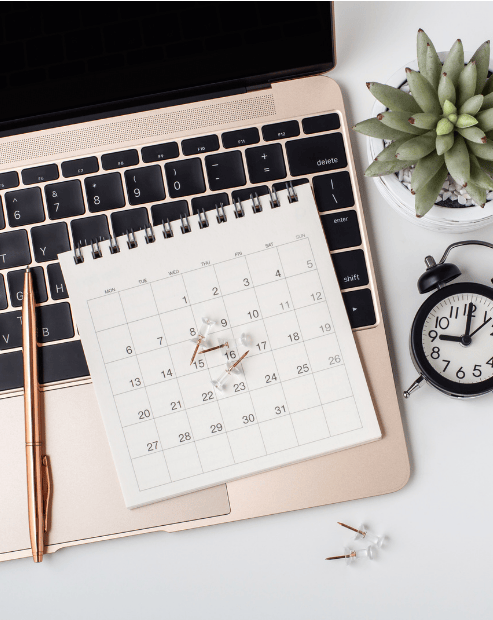 Small hero image promoting the Future Planning for Creatives workshop. A rose-gold laptop is partially open, with a spiral-bound monthly calendar resting directly on its keyboard. Several clear thumbtacks are scattered on the calendar and the desk nearby. Surrounding the laptop are a matching rose-gold pen, a small black vintage-style alarm clock, and a succulent in a white pot. The overall aesthetic is organized, professional, and focused on productivity and scheduling.