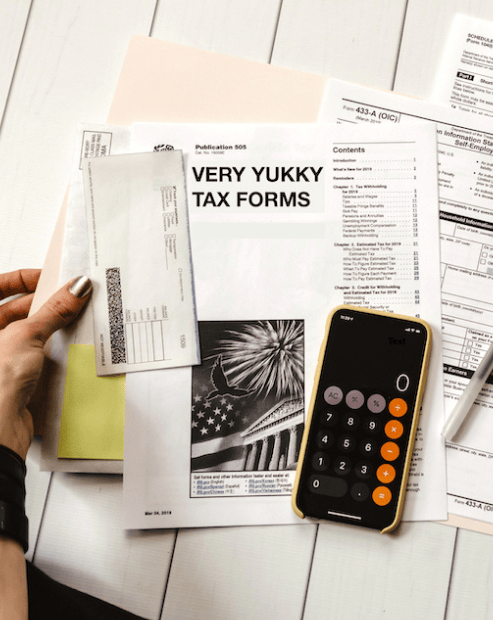 A top-down view of a desk with several official-looking documents, including one titled "VERY YUKKY TAX FORMS". A hand with metallic nail polish holds an envelope over a folder, while a smartphone displaying a calculator app sits next to a pen on a white wooden surface.