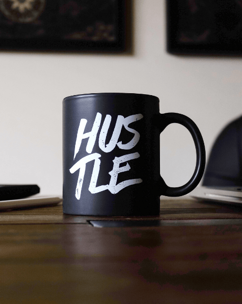 Small promo image for  The Creative Hustle Workshop. A close-up shot of a matte black coffee mug sitting on a wooden desk. The mug features the word "HUSTLE" in bold, white, brush-style lettering.