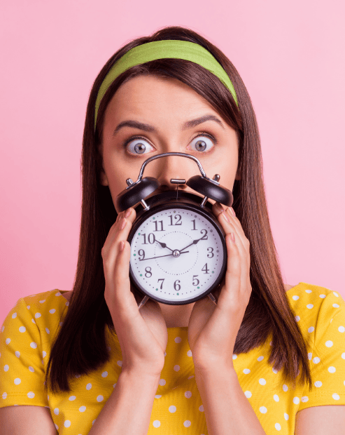 Small hero image for the time management for creatives workshop. A woman with a startled expression wearing a yellow polka-dot shirt and green headband, holding a black vintage alarm clock in front of her face against a bright pink background.