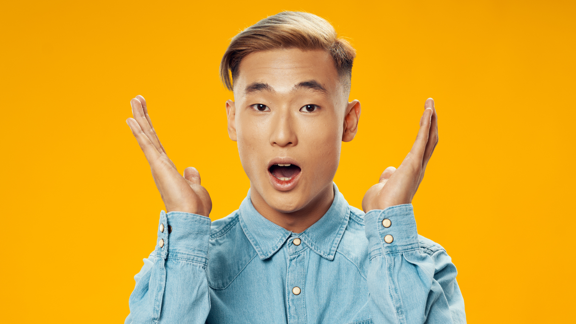 Blog April 2026 – What assessors look for in a arts grant application A young East Asian man with short, styled brown hair and a faded undercut stands against a solid, vibrant yellow background. He wears a light-wash denim button-down shirt. His expression is one of exaggerated surprise or amazement, with his mouth open and his hands raised palms-forward near his face. The lighting is bright and even, highlighting his features and the texture of the denim.