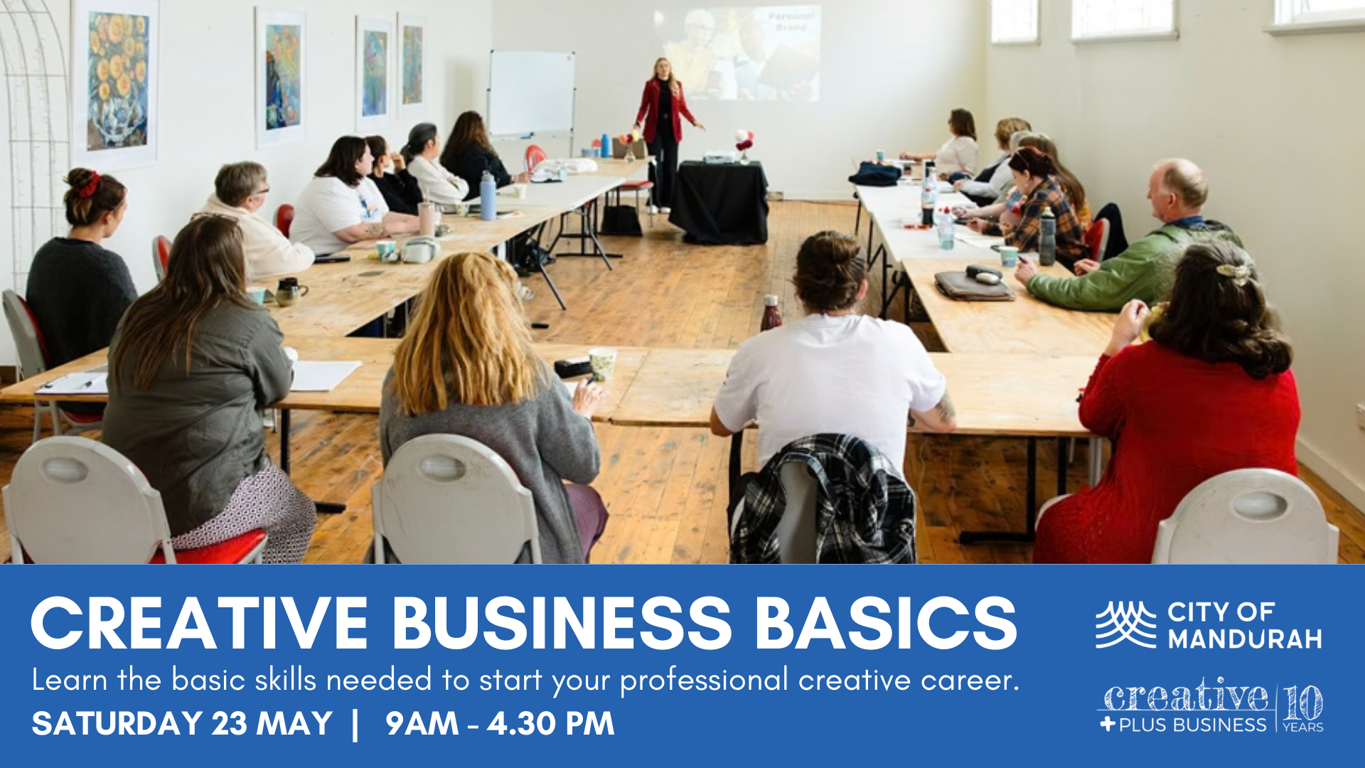 A promotional banner for a "Creative Business Basics" workshop. The top half shows a wide-angle photo of a workshop in progress: a diverse group of people sits around a U-shaped wooden table in a brightly lit room with artwork on the walls. A presenter in a red blazer stands at the head of the table. The bottom half features a solid blue footer with white text: "CREATIVE BUSINESS BASICS: Learn the basic skills needed to start your professional creative career. Saturday 23 May | 9am – 4:30 pm." Logos for City of Mandurah and Creative Plus Business are in the bottom right corner.