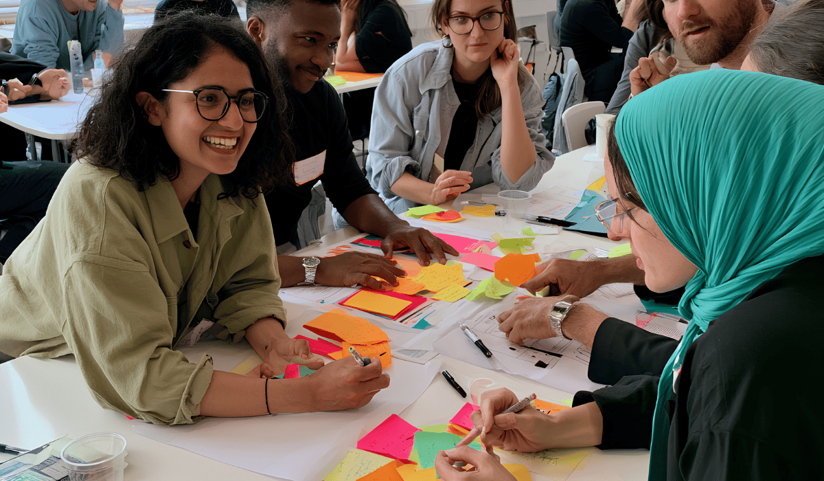 Hero image for the Creative Plus Business Programs page, featuring a vibrant, diverse group of participants engaged in a hands-on professional development workshop. People are seated around a white table filled with bright sticky notes, markers, and worksheets, smiling and collaborating. The page provides information on long-form and full-day training programs for creative industries and communities.